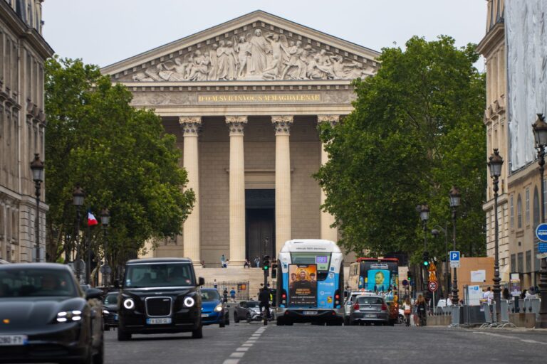 A vibrant Paris street scene with vehicles passing in front of the iconic La Madeleine church.